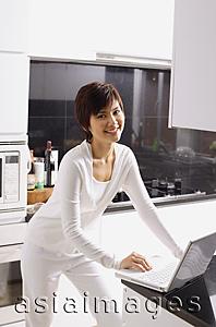 Asia Images Group - Young woman standing in kitchen, using laptop, smiling at camera