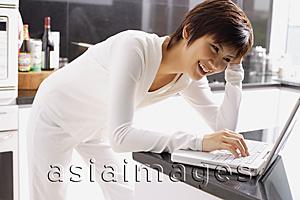 Asia Images Group - Young woman leaning on kitchen counter, using laptop