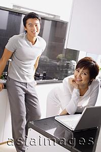 Asia Images Group - Couple in kitchen, woman leaning on kitchen counter with laptop open in front of her