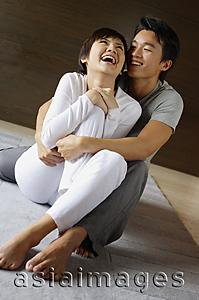 Asia Images Group - Couple sitting on floor, man embracing woman from behind, woman laughing