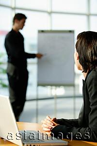 Asia Images Group - Female executive sitting at desk, turning to look at colleague standing next to flip board