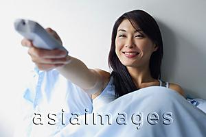 Asia Images Group - Woman sitting up in bed, holding TV remote control, arm outstretched