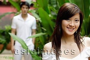 Asia Images Group - Young woman in garden, smiling at camera, man in the background