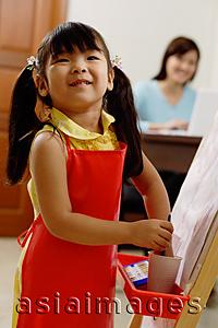 Asia Images Group - Young girl standing next to easel, smiling at camera, mother in the background