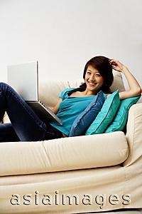 Asia Images Group - Woman reclining on sofa with laptop, looking at camera