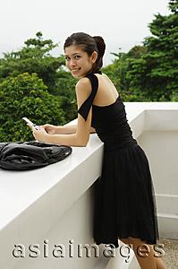 Asia Images Group - Young woman in black dress, leaning on ledge, holding mobile phone