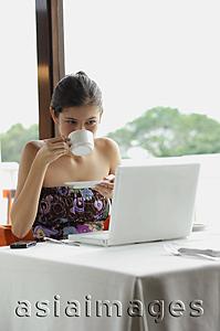 Asia Images Group - Woman sitting in restaurant, using laptop, drinking from cup