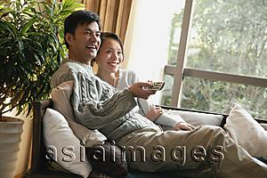Asia Images Group - Couple in living room, sitting side by side on sofa, man holding TV remote control