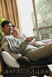 Asia Images Group - Couple sitting side by side on sofa, man holding TV remote control