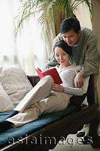 Asia Images Group - Couple in living room, woman reading a book, man leaning over her