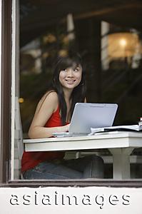 Asia Images Group - Young woman sitting at table, using laptop, looking through window