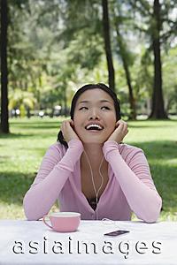 Asia Images Group - Young woman sitting outdoors, listening to MP3 player
