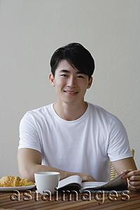 Asia Images Group - Young man having breakfast and reading a magazine, smiling at camera