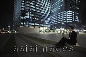 Asia Images Group - Man sitting on ground working on lap top in front of lit buildings