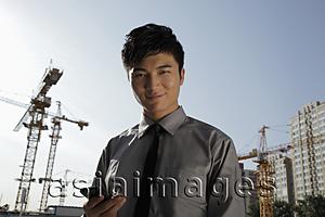 Asia Images Group - Young man holding phone standing in front of construction site