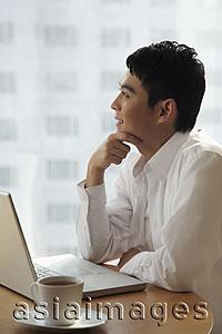 Asia Images Group - Young man sitting with laptop looking out window