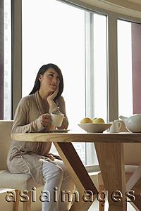 Asia Images Group - Young woman sitting at table holding a cup and looking up