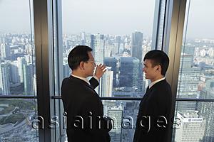 Asia Images Group - Two men talking in front of window with a view of the city of Beijing