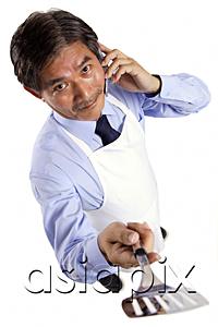 AsiaPix - Mature man wearing apron, using mobile phone, holding spatula towards camera