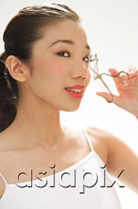 AsiaPix - Woman holding eyelash curler to eye, looking at camera