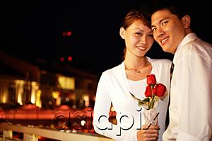 AsiaPix - Couple standing side by side, woman holding single rose stalk, both looking at camera