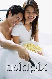 AsiaPix - Couple sitting on bed, smiling, watching TV, man leaning on womans shoulder