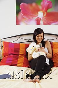 AsiaPix - Girl sitting on bed, hugging stuffed toy, using mobile phone