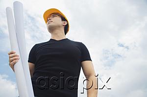 AsiaPix - Young man wearing hardhat, carrying blueprints, outdoors