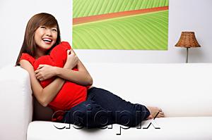 AsiaPix - Young woman sitting on sofa, embracing pillow