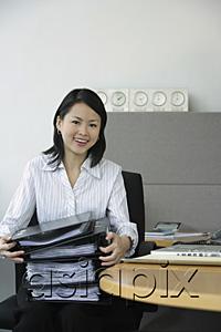 AsiaPix - Young woman sitting at office desk, holding stack of binders