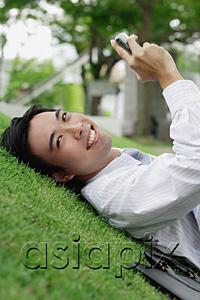 AsiaPix - Businessman lying on grass in park, holding mobile phone, smiling at camera