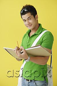 AsiaPix - Man standing against yellow background, writing in notebook, looking at camera