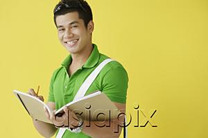 AsiaPix - Young man writing in notebook, looking at camera