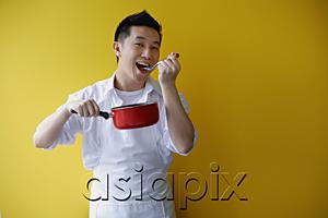 AsiaPix - Man wearing apron and holding saucepan, eating from spoon