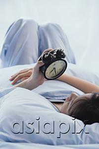 AsiaPix - Young woman lying on bed, looking at alarm clock