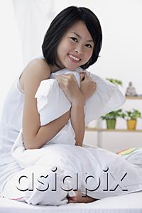AsiaPix - Young woman sitting on bed, hugging pillow, smiling