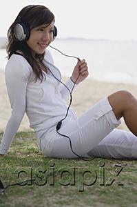 AsiaPix - Young woman sitting on beach, listening to music, wearing headphones