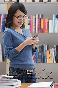 AsiaPix - Young woman in library, using mobile phone, looking at camera