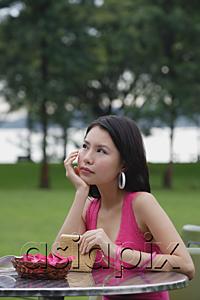 AsiaPix - Young woman sitting at outdoor table, hand on chin, looking away