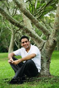 PictureIndia - Young man sitting under tree, listening to music, looking at camera