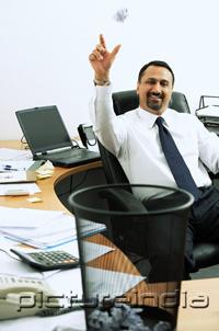 PictureIndia - Businessman sitting in office throwing paper ball into dustbin, smiling