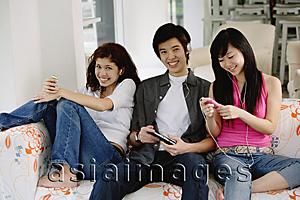 Asia Images Group - Teenagers sitting side by side, on sofa, listening to music from MP3 players